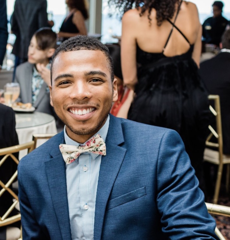Bryon Lewis-Cummings seated at a formal event in a blue suit with a floral bow tie